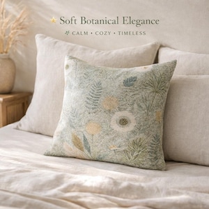 May include: A square cushion with a botanical print in shades of green, cream, and yellow, featuring flowers and foliage. The cushion is on a bed with neutral-coloured bedding. The text "Soft Botanical Elegance" is visible.