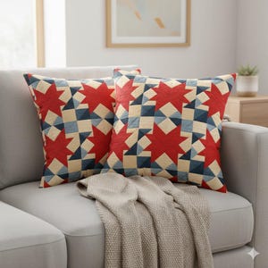 May include: Two decorative throw pillows with a patchwork design featuring red starbursts and geometric patterns in blue, cream, and navy. The pillows rest on a gray sofa with a beige throw blanket.