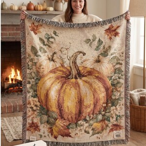 May include: A woven throw blanket featuring a large central orange pumpkin surrounded by smaller pumpkins, autumn leaves, and greenery on a cream-colored background. The blanket has a fringed edge and a rectangular tag in the bottom corner.