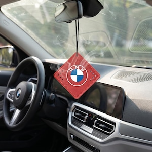 May include: A square red leather car air freshener hanging from the rearview mirror. The air freshener features a stitched border and the circular BMW logo in the centre with blue, white, and black colours. A thin black cord attaches it to the mirror.