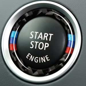 Può includere: Primo piano del pulsante di avviamento/arresto del motore di un'auto. Il pulsante nero ha il testo bianco "START STOP ENGINE". Il pulsante è circondato da un anello argentato e da un motivo in fibra di carbonio. Una striscia blu, bianca e rossa è sul lato.