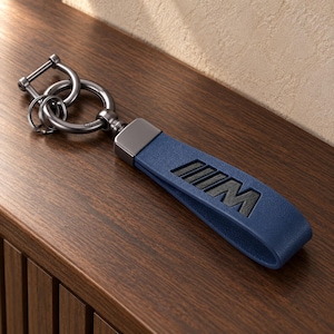 May include: A dark blue leather keychain featuring the embossed black M logo. The keychain includes a gunmetal gray metal D-ring, a circular ring, and a smaller split ring for keys, all connected by a matching dark gray metal clasp.