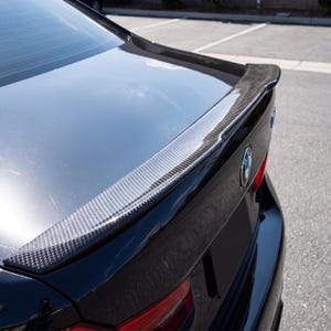 Op de afbeelding: Een carbon fiber achterspoiler gemonteerd op een zwarte auto. De spoiler is donkergrijs met een geweven patroon. De achterklep en achterruit van de auto zijn zichtbaar en weerspiegelen de lucht. Het BMW-embleem is zichtbaar.