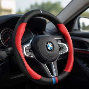 Op de afbeelding: Een zwart en rode stuurwielhoes met een BMW-logo. De hoes heeft rode geperforeerde leren secties en een zwarte boven- en onderkant. Een blauw-rode streep is zichtbaar aan de onderkant van het stuur.
