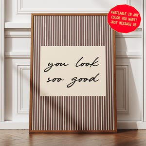 Peut inclure: Une affiche encadrée rectangulaire avec un fond à rayures verticales marron et blanc et un rectangle central crème portant le texte noir en cursive "you look soo good". Un badge circulaire rouge est visible dans le coin.