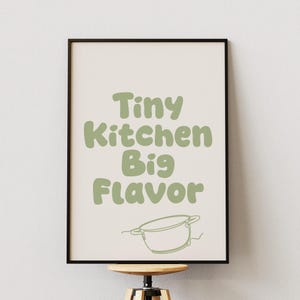 Peut inclure: Une affiche encadrée avec le texte "Tiny Kitchen Big Flavor" en vert, sur fond crème, avec un dessin d'une casserole. Le cadre est noir, parfait pour la décoration de cuisine.