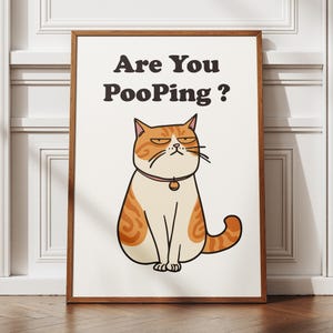 Peut inclure: Une affiche murale rectangulaire dans un cadre en bois marron représentant un chat tigré orange et blanc de style dessin animé avec une expression grincheuse. Le chat porte un collier rose avec une clochette. Le texte indique : Are You PooPing ?