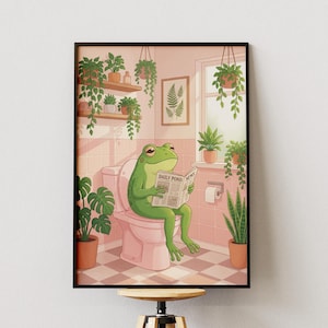 Peut inclure: Une impression d'art encadrée représente une grenouille verte assise sur des toilettes roses, lisant un journal intitulé "Daily Pond News". La salle de bain comprend des plantes en pot, une fenêtre et un mur carrelé rose. L'art a un style illustratif fantaisiste.