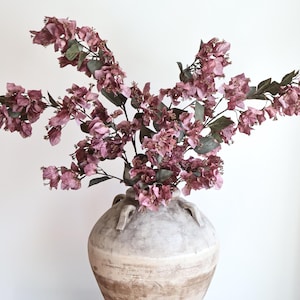 May include: A large, weathered, light-coloured ceramic amphora vase with two small handles, containing a sprawling arrangement of artificial bougainvillea branches featuring dusty mauve-pink paper-like bracts and dark green leaves.