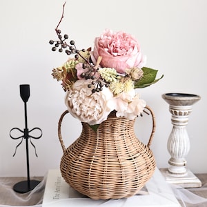 Könnte beinhalten: Eine gewebte Korbvase gefüllt mit rosa und cremefarbenen Blumen und Grün. Die Vase hat zwei Griffe und steht auf einem weißen Buch mit dem Titel "The Art of Home". Ein schwarzer Metallkerzenhalter und ein weißer Kerzenhalter befinden sich auf beiden Seiten.