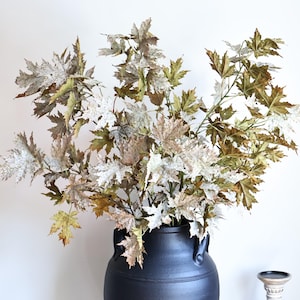 May include: A large black ceramic vase with two side handles containing a cluster of artificial maple branches. The leaves feature a mix of muted green, brown, and white speckled tones. A small white wooden candle holder stands to the right.