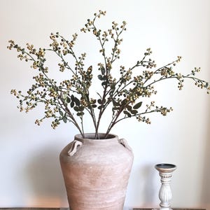 May include: A large, light brown terracotta vase with two small side handles, containing several branches with small, round, metallic gold and green berries and dark green leaves, positioned next to a small, white, fluted pedestal candle holder.