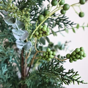 May include: An artificial floral arrangement featuring dark green cedar-like foliage, light green berry clusters on thin stems, and pale, dusty green eucalyptus leaves with a matte texture, all combined into a single botanical spray.