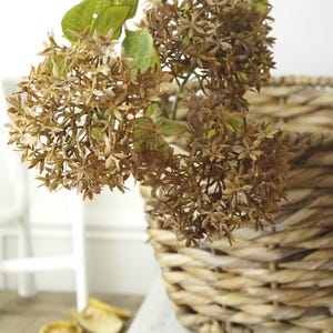 May include: A woven basket filled with dried brown flowers and green leaves. The basket sits on a wooden surface next to a book titled "The Art of Home". The overall aesthetic is rustic and natural.