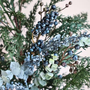 May include: A decorative floral arrangement featuring dark green cedar-like foliage, clusters of small dark blue berries with a frosted finish, and pale green eucalyptus leaves on thin brown stems, creating a textured botanical display.