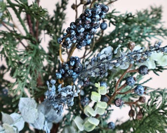 Peut inclure: Un arrangement floral décoratif composé de feuillage vert foncé rappelant le cèdre, de grappes de petites baies bleu foncé à la finition givrée et de feuilles d'eucalyptus vert pâle sur de fines tiges brunes texturées.
