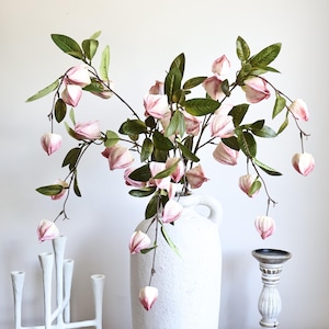 May include: A tall, white, textured ceramic jug holds several branches with green leaves and pale pink, bell-shaped flowers. To the left is a white, multi-armed candle holder, and to the right is a small, white, fluted wooden candle pedestal.