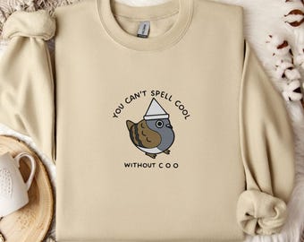 May include: A beige sweatshirt with a round neck and long sleeves. The front features a cartoon pigeon wearing a hat, with the text "You can't spell cool without coo" in a circular design. The sweatshirt is displayed on a textured surface.