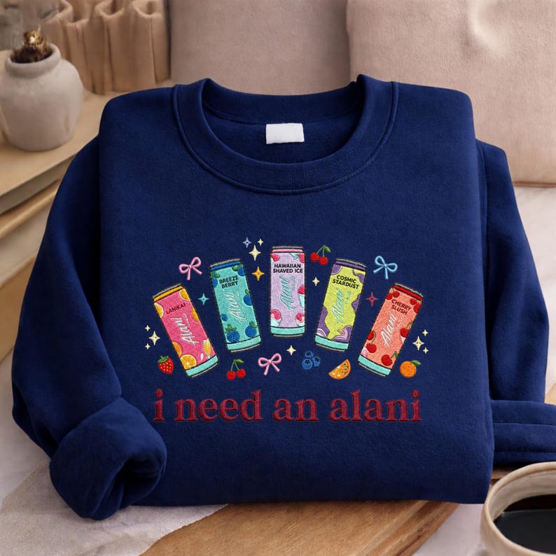 A navy blue sweatshirt with a colorful graphic design featuring five cans of soda and the text "I need an alani" printed on it.
