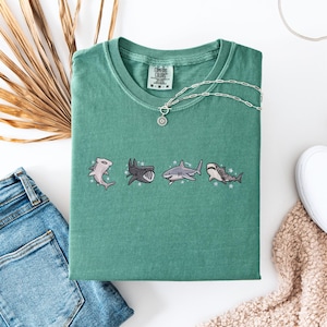 a green t-shirt with a graphic of four cartoon sharks, a pair of blue jeans, a pair of white sneakers, and a beige sweater.