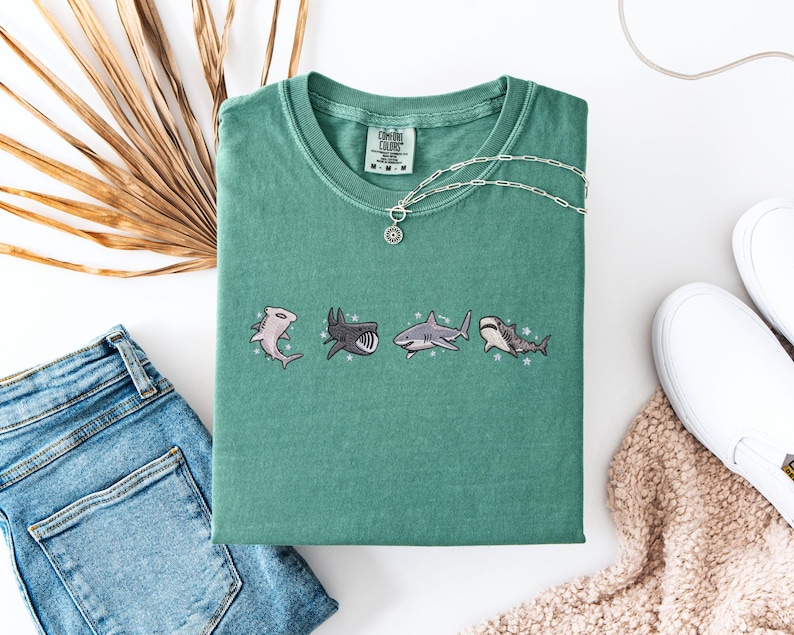 a green t-shirt with a graphic of four cartoon sharks, a pair of blue jeans, a pair of white sneakers, and a beige sweater.