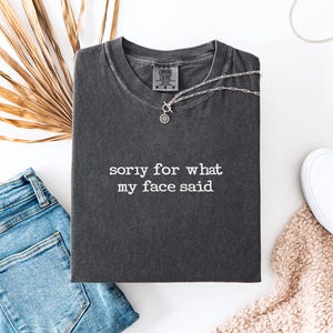 May include: Dark gray t-shirt with the text "sorry for what my face said." The shirt is folded and has a silver necklace on top. A pair of blue jeans, white sneakers, and a beige blanket are also in the image.