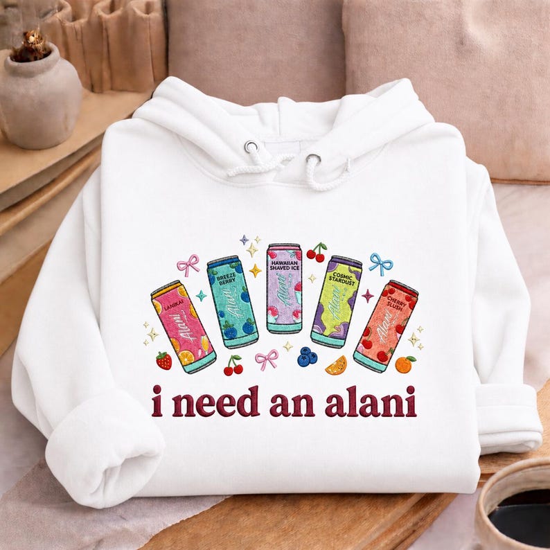 A white hoodie with a graphic design featuring five colorful cans of soda and various fruits, along with the text "I need an alani" printed on it.