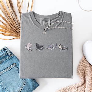 a casual outfit consisting of a gray t-shirt with shark designs, a pair of blue jeans, a necklace, and a pair of white sneakers.