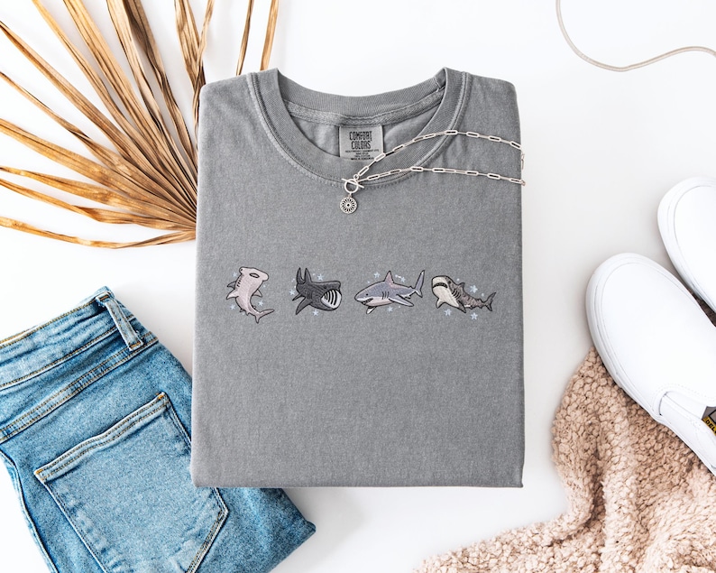 a casual outfit consisting of a gray t-shirt with shark designs, a pair of blue jeans, a necklace, and a pair of white sneakers.