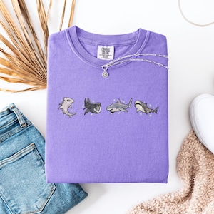 a purple t-shirt with a graphic of four cartoon sharks, a pair of blue jeans, a pair of white sneakers, and a beige sweater.