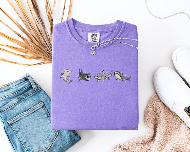 a purple t-shirt with a graphic of four cartoon sharks, a pair of blue jeans, a pair of white sneakers, and a beige sweater.