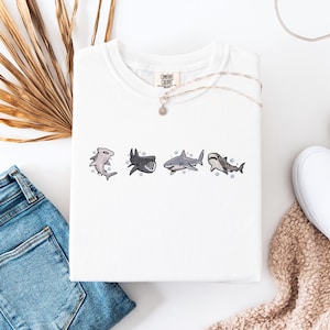 a white t-shirt with a graphic of four sharks, a pair of blue jeans, a pair of white sneakers, and some dried leaves or straw.
