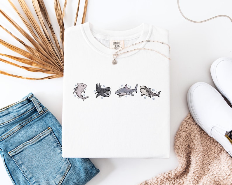 a white t-shirt with a graphic of four sharks, a pair of blue jeans, a pair of white sneakers, and some dried leaves or straw.
