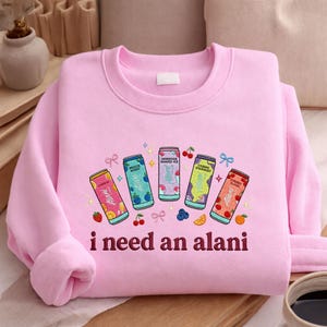 A pink sweatshirt with a graphic design featuring five colorful cans of soda and the text "I need an alani" printed on it.