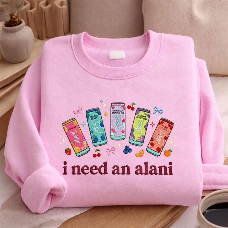 A pink sweatshirt with a graphic design featuring five colorful cans of soda and the text "I need an alani" printed on it.
