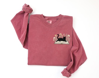 May include: A dusty rose long-sleeve sweatshirt with a crew neck. The front features an embroidered design of a black cat lying on an open book, surrounded by flowers. The brand name "COMFORT COLORS" is on a small tag.