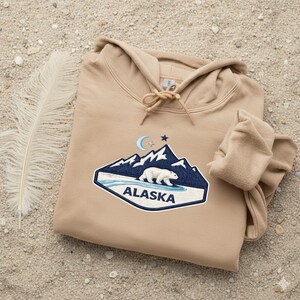 May include: A tan hoodie with a drawstring hood and a front pocket. The hoodie features an embroidered patch with a polar bear, mountains, and the word "Alaska". The patch is blue, white, and tan.
