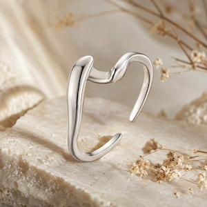 May include: A silver-toned open ring featuring a smooth, fluid, and wavy design. The metal band has a polished, reflective surface with an irregular, organic shape that curves upwards and downwards, creating an open gap for adjustable wear.
