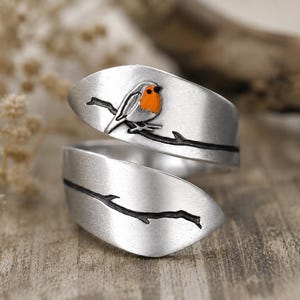 May include: A silver wrap-around ring with a detailed design of a robin perched on a branch. The bird has an orange breast and is set against a brushed metal finish. The ring's design wraps around the finger.