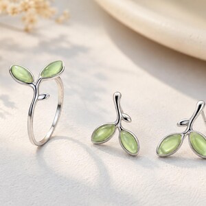 May include: A silver ring and a pair of matching stud earrings, each featuring a branch design with two small, smooth, light green oval stones shaped like leaves. The metal has a polished finish, and the stones have a soft, translucent appearance.