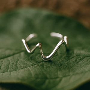 Peut inclure: Une bague argentée présentant un design ondulé ou en zigzag continu et lisse. L'anneau est ouvert sur le dessus, créant un espace entre les deux extrémités du fil métallique. La surface de la bague est polie et réfléchissante, captant la lumière.