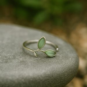 Op de afbeelding: Een zilveren ring met een delicaat bladerontwerp. De ring heeft twee lichtgroene, ovaalvormige stenen, die op bladeren lijken, op een slanke band. De ring wordt getoond op een grijze steen, met een vage groene achtergrond.