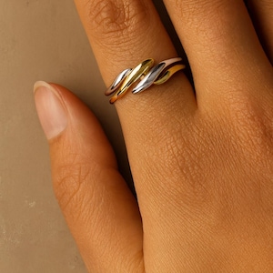 May include: A tri-colour metal ring featuring three intertwined, smooth, polished bands in yellow gold, rose gold, and silver tones. The bands are layered and curved, creating a continuous, flowing design that wraps around the finger.