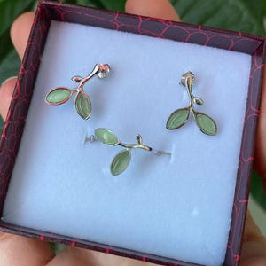 May include: A jewelry set consisting of two stud earrings and one ring, all featuring a silver-toned metal branch design with small, smooth, light green oval-shaped leaves. The items are displayed inside a dark red box with a white interior.