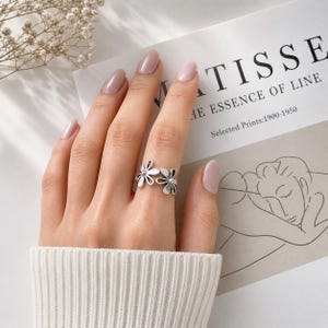 Peut inclure: Une bague argentée composée de deux motifs de fleurs de marguerite ajourées reliés par un anneau fin. Les pétales sont allongés et arrondis, avec un petit point circulaire au centre de chaque fleur. La bague est portée sur un doigt.