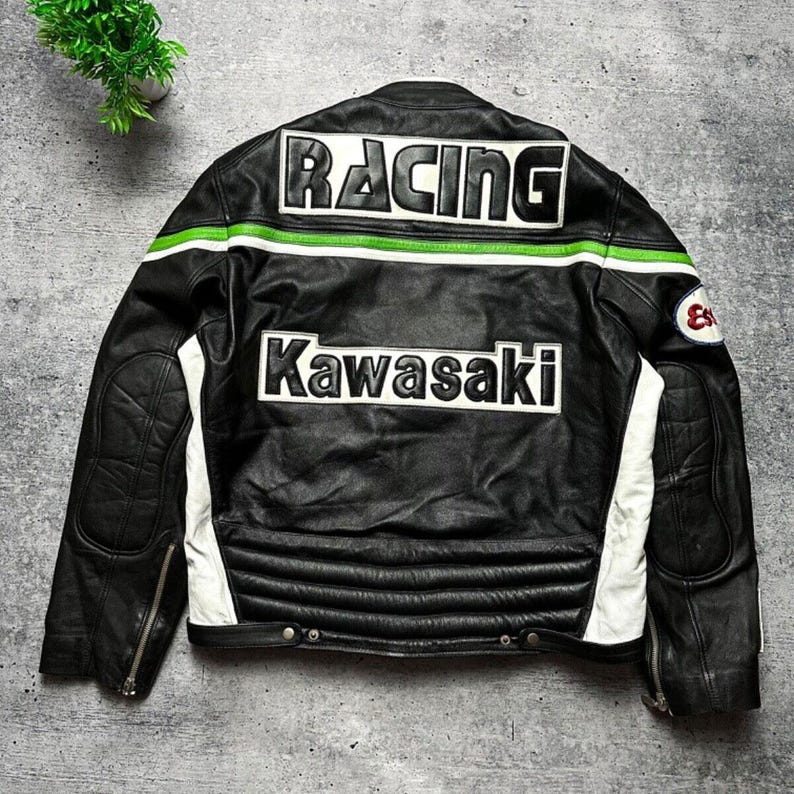 May include: Black leather racing jacket with white and green accents. The back features the word "RACING" across the shoulders and "Kawasaki" below. The jacket has a quilted design on the lower back and white panels on the sides.