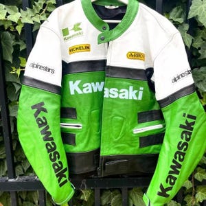 Pode incluir: Casaco de corrida Kawasaki vintage, branco, verde e preto. Apresenta logótipos da Kawasaki, com mangas verdes com a inscrição "Kawasaki". Tem gola alta e vários patches, refletindo uma estética de corrida clássica.