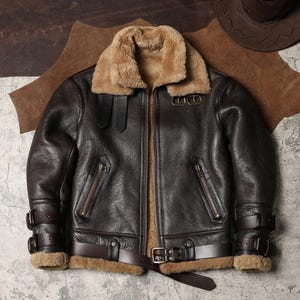 Puede incluir: Chaqueta de cuero marrón oscuro con cuello y forro de borrego. La chaqueta tiene cierre de cremallera, múltiples bolsillos y correas ajustables con hebillas. La chaqueta tiene un estilo aviador clásico.
