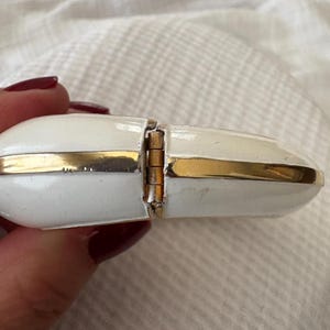 May include: A white and gold hinged jewelry box. The box is oval-shaped with a gold band around the edges. The hinge is visible, and the box appears to be made of enamel. The background is a white, textured surface.