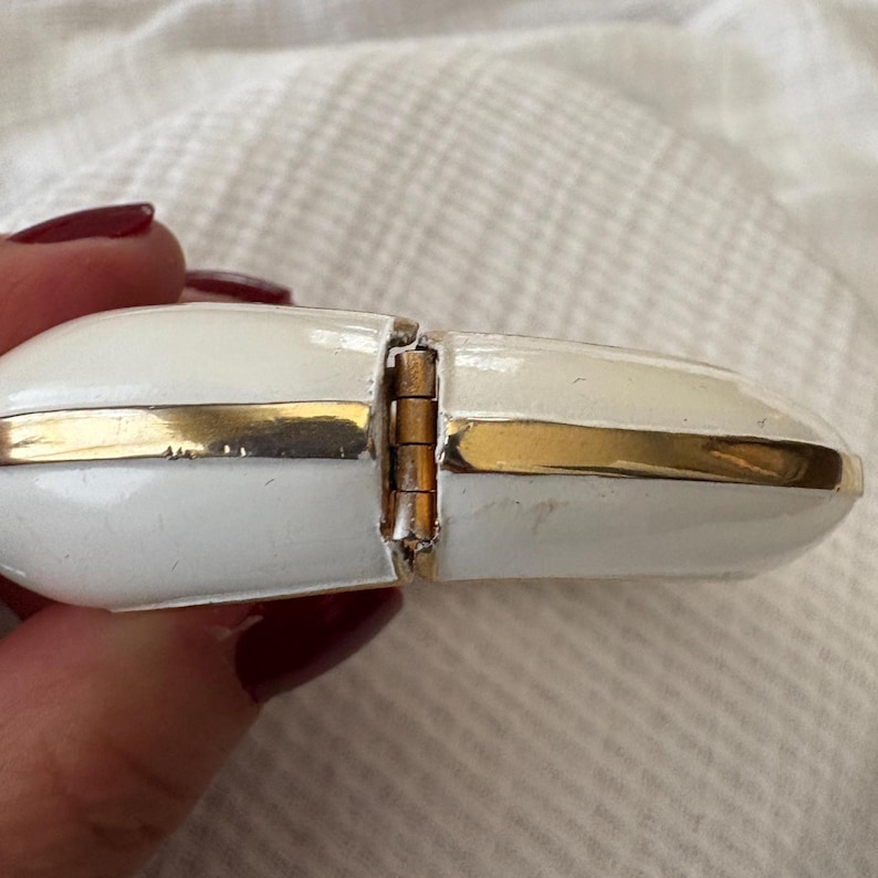 May include: A white and gold hinged jewelry box. The box is oval-shaped with a gold band around the edges. The hinge is visible, and the box appears to be made of enamel. The background is a white, textured surface.